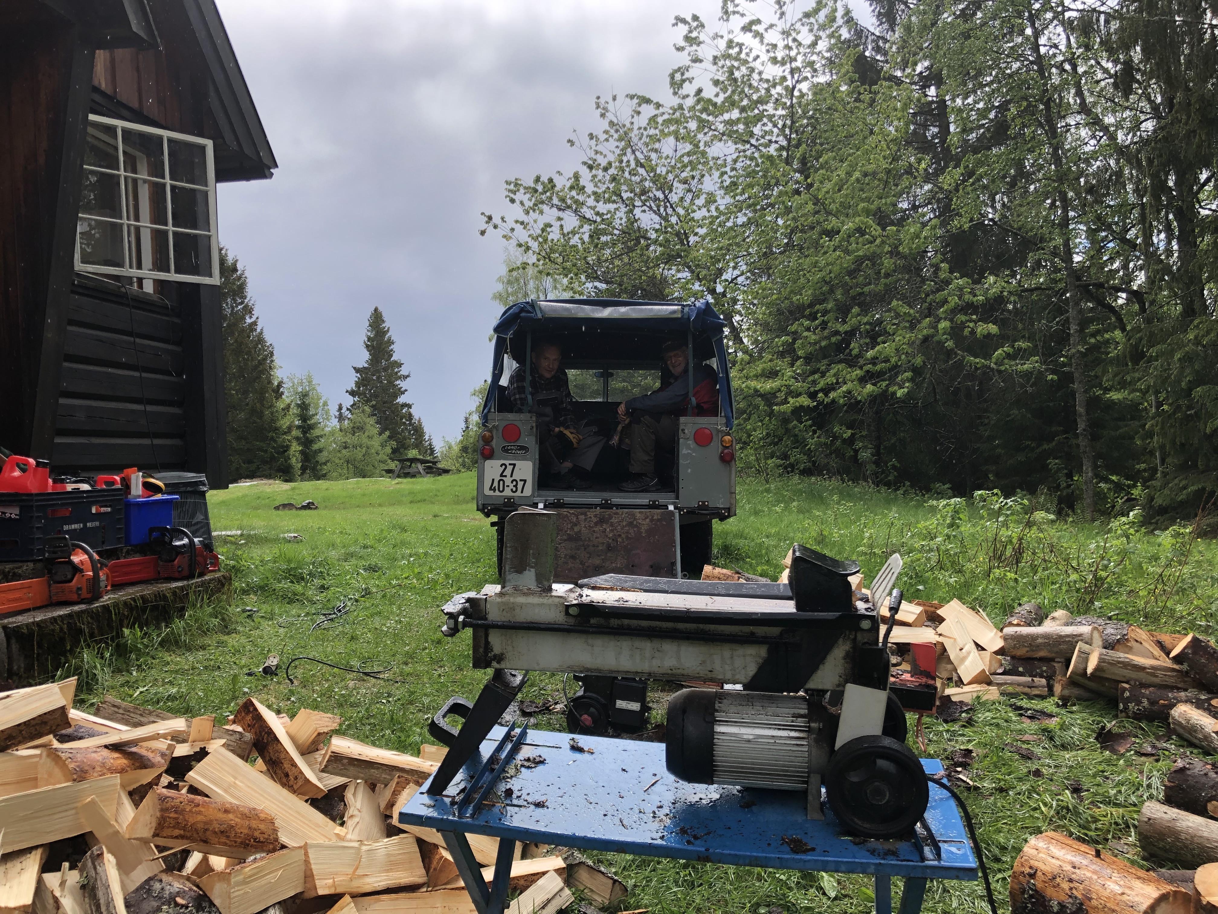 Vedkløyver med ved omrking og bil med bakdør åpen. Dugnadsgjeng i bil. Foto.